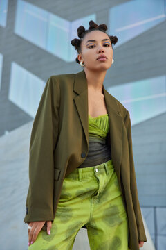 Self confident fashionable teenage girl dressed in green jacket and jeans has funny hairstyle poses against grey wall in public place has serious expression. Youth fashion and style concept.