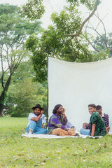 Malaysian Indian men in a group against a cloth backdrop in a park surrounded by trees, talking,...