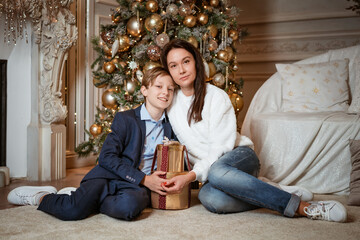 Mother and son are sitting on the floor in the room in front of a Christmas tree with gift boxes in their hands. They smile.