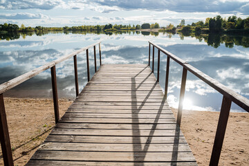 Panorama of the pier by a quiet lake on a sunny day. A wooden pier at the water surface, clouds are reflected in the water, and a forest grows around.