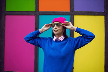 fashionable brunette woman in a pink warm hat and sweater and sunglasses stands against the background of colorful windows in autumn
