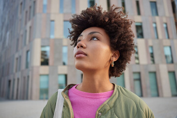 Close up shot of thoughtful curly haired woman focused above thinks about something poses outdoors against modern building background walks in city during free time goes sightseeing explores place