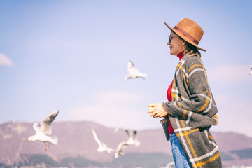 Young woman traveler in hat standing on the beach feeding seagulls