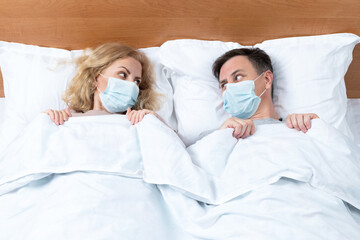 Couple in bed, man and woman in respiratory mask lie under the covers and look at each other. Relationships in a pandemic.