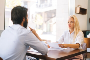 Side view of unrecognizable business man and attractive buisinesswoman sitting in cafe and discussing contract. Diverse businesspeople meeting in hotel lobby reading documents.