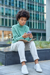 Fototapeta premium Serious curly woman in casual clothes and white sneakers has opened notebook on knees checks newsfeed on smartphone sits outdoors against modern glass city building. Urban lifestyle concept.