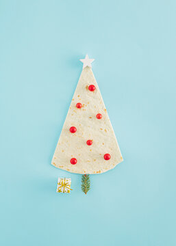 Creative Christmas tree idea made of a tortilla or pizza dough, a white wooden star, pine twig and red baubles on a blue background. Minimal flat lay, New Year concept. Christmas food inspiration.