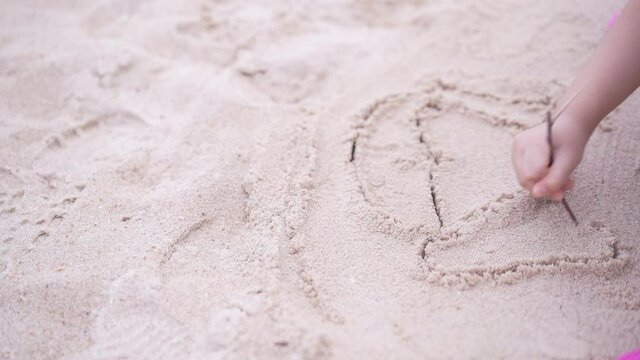 closeup hand of asian child or kid girl drawing woman smiling face picture on sandy beach by twigs on family holiday vacation at sea on slow motion with copyspace