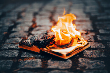 An open old book lies on the sidewalk and burns with a bright flame. Book burning and censorship. The Antichrist. - obrazy, fototapety, plakaty