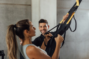 Smiling male Coach helps woman with suspension strap workout at gym hiit class.