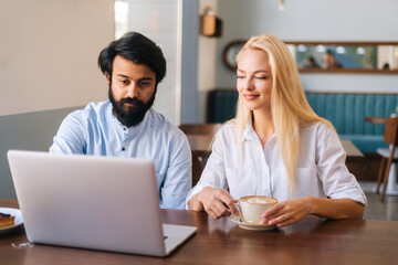 Front view of two business people of bearded Indian man and attractive Caucasian blonde woman working with laptop in cafe and drinking coffee. Friendly colleagues discussing project in coffee shop