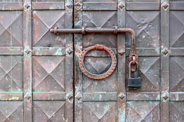 An old wrought iron door knocker, a metal latch and a lock on an old metal door in the Russian style.  The forged hammer handle and the handmade latch are made of iron in close-up against the backgrou