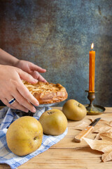 Close-up of woman's hands holding an apple pie on wooden table with pippin apples and candle, vertical