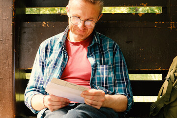 A young man in glasses and a shirt is reading a letter while sitting in the park in the gazebo....