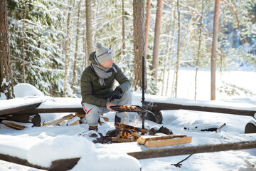 Man roasting sausages on campfire in forest by the lake, making a fire, grilling. Happy tourist exploring Finland. Beautiful sunny winter landscape, wood covered with snow. 