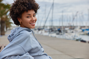 Horizontal shot of cheerful woman with curly hair wears blue hoodie looks happily at camera poses outside at harbor feels relaxed admires scenic views. People recreation and lifestyle concept