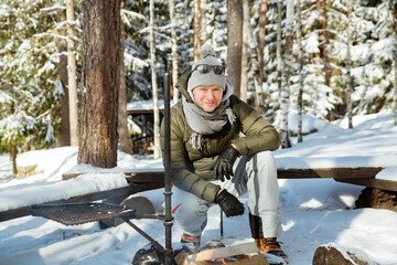 Fototapeta na wymiar Man roasting sausages on campfire in forest by the lake, making a fire, grilling. Happy tourist exploring Finland. Beautiful sunny winter landscape, wood covered with snow. 