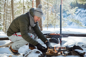 Man roasting sausages on campfire in forest by the lake, making a fire, grilling. Happy tourist exploring Finland. Beautiful sunny winter landscape, wood covered with snow. 