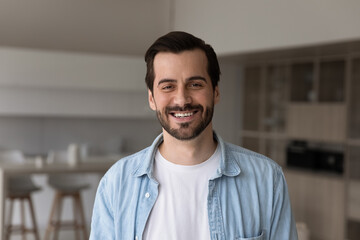 Head shot portrait of young smiling handsome man feeling satisfied with dental services, posing in modern living room. Happy millennial gen male homeowner looking at camera, standing in own flat.
