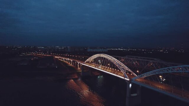 A motorcade of three cars rides over a bridge over a river at night