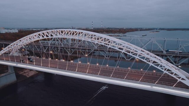 A motorcade of three cars drives across the bridge over the river at dawn, a boat sails under the bridge