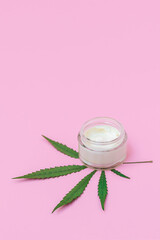 Cosmetic glass jar with cannabis-infused face cream and green marijuana leaf on pastel pink background, vertical image