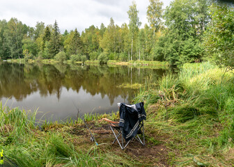 Fototapeta na wymiar lake shore and fisherman's chair, calm lake water surface, tree reflections, cloudy day, fishing concept
