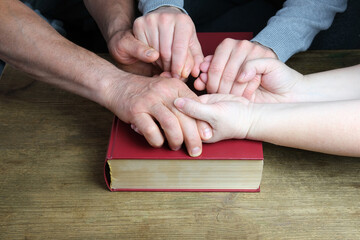 close-up of the hands of two men and a woman folded in a prayer gesture on a thick family bible in...