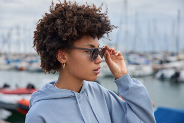 Young curly female tourist wears sunglasses and sweatshirt focused into distance strolls at...