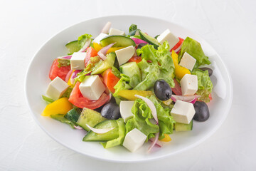 Greek salad of fresh cucumber, tomatoes, bell peppers, lettuce, red onions, feta cheese and olives with olive oil in a white plate on a light background. Healthy eating. Close up