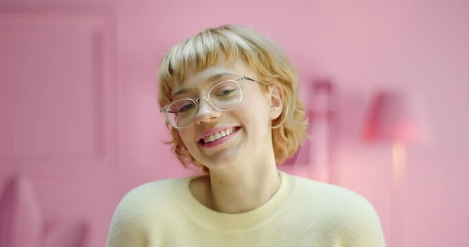 Studio shot of a cute young girl staring at the camera wearing glasses and smiling with ashamed expression on a pink color studio background. Happy Caucasian girl with blonde hair showing smile