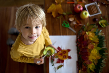 Sweet child, boy, eating avocado while applying leaves using glue while doing arts and chraft with leaves