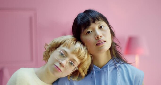 Portrait of two romantic diversity lesbian couple leaning heads each other looking at the camera in pink studio. Young happy multiracial women friends standing together and lean heads each other