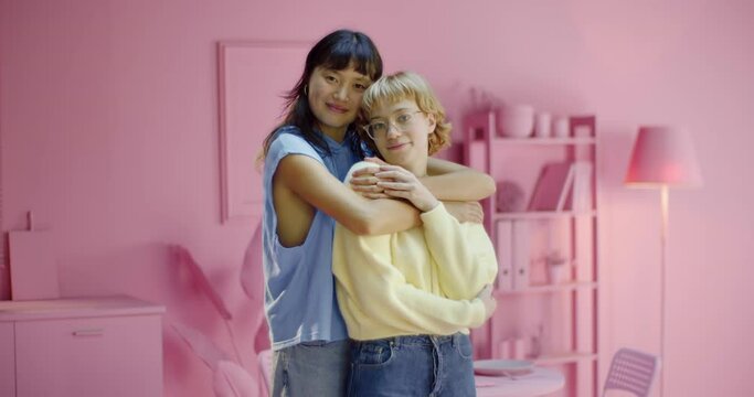 Portrait of pretty lesbian woman embracing her girlfriend smiling and looking at the camera standing at pink trendy studio. Multiracial female couple hugging enjoying happiness at home