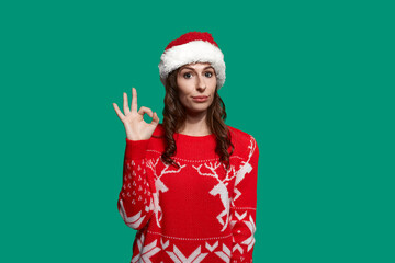 Calm woman with dark hair shows ok gesture, says okay, approves quality of a product, recommends service, wears red ugly sweater with deer and santa hat on green background. Christmas concept.