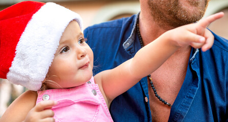 merry christmas! young baby girl portrait celebrating xmas wearing a santa hat outdoor sitting on dad knees. father and child close up during winter warm festivities. family time on summer vacation