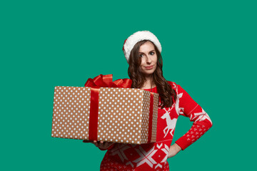 Pretty european woman with dark hair holds a big gift box with bow knot, getting ready to give presents, wears red ugly sweater with deer and santa hat on green background. Christmas concept.