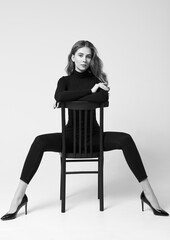 Studio portrait of beautiful skinny girl. Pretty young woman dressed in turtleneck sweater, leggings and shoes sitting on stool. Black and white