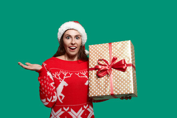 Happy inspired woman is holding big gift box with bow knot and throws palm aside, has eyes and mouth wide open, wears red ugly sweater with deer and santa hat on green background. Christmas concept.