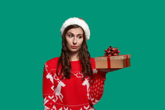 Frowny brunette holds golden gift box with bow knot on palm and looks suspiciously at it, wants other present, wears red ugly sweater with deer and santa hat on green background. Christmas concept.