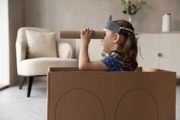 Side view adorable little preschool child girl sitting in huge cardboard box, playing pirates game alone at home. Playful smiling small cute kid daughter involved in entertaining domestic activity.