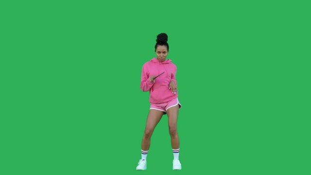 Young woman with black curly hair dancing over green background. African american girl table tennis player wearing pink hoodie and shorts having fun with racket. Chroma key. Copy space