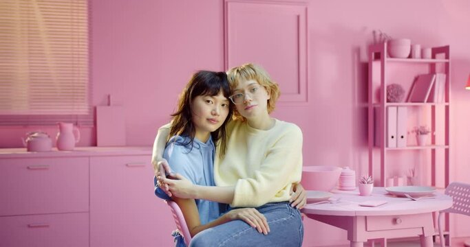 Portrait of young Caucasian lesbian woman sitting on her partner lap looking at camera enjoying togetherness at pink color studio background. Multiracial female couple sitting on partner lap at home