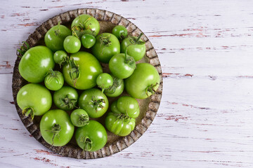 Green tomatoes on a brown plate, on a white wooden table, top view.