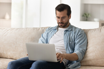 Concentrated young man in eyewear sitting on couch with laptop, involved in typing message or...