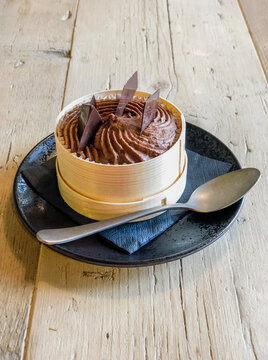 Présentation d'un dessert de mousse au chocolat et copeaux de chocolat dans une assiette noir présenté dans un cerclage de bois sur une table en bois
