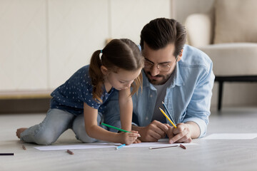 Happy adorable kid girl drawing pictures on paper sheet with caring young father, lying together on heated wooden floor. Smiling bonding different generations family involved in creative activity.