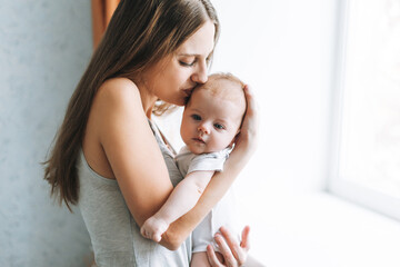 Young woman mom with long hair holding baby girl on hands near window at home