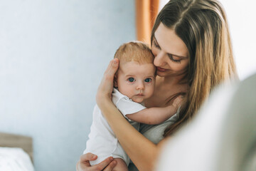Young woman mom with long hair holding baby girl on hands near window at home