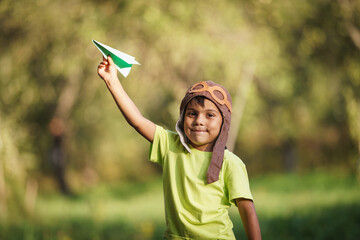 cute little Latin American boy in a pilot's hat plays as a pilot, launching a paper airplane into air. child is happy about the flight of the airplane. Children's active outdoor game. Dynamic image